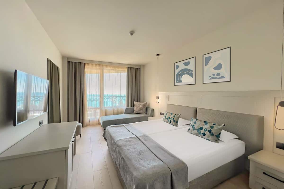 Habitación de hotel con una cama grande, un escritorio, una silla, un televisor y un balcón con cortinas.