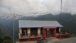 Un edificio pequeño con techo de metal y base de ladrillo, situado en una zona montañosa con una carretera en primer plano y vistas de las montañas al fondo.