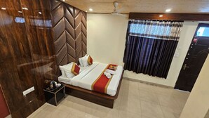 Una cama bien hecha con almohadas y una colcha roja y dorada, un cabecero de madera, una mesita de noche con una tetera, y una ventana con cortinas estampadas.