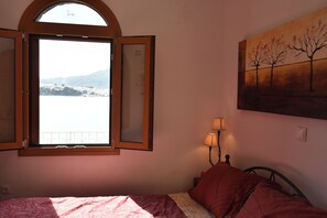 A bedroom with a bed, a window with an open shutter, a painting on the wall, and a lamp on the nightstand. - Porto Castello Villas House of Zisis (Skiathos)