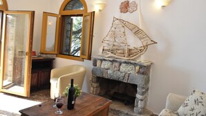 Ein gemütliches Wohnzimmer mit Kamin aus Stein, einem Holztisch mit einer Weinflasche, einem beigen Sessel und einem großen Fenster mit geöffneter Tür.