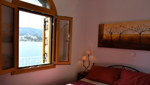 Ein Schlafzimmer mit Bett, Nachttisch, Fenster mit Blick auf ein Gewässer und Berge sowie einem Bild an der Wand.