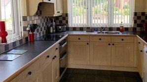 Uma cozinha com armários de madeira clara, bancada preta e painel de azulejos. Há uma janela com cortinas brancas e uma chaleira vermelha no balcão.