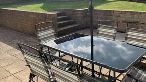Um pátio externo com mesa e cadeiras, um grande guarda-sol e um casebre de madeira ao fundo.