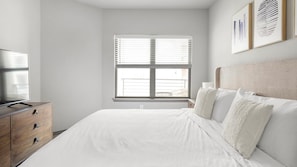 A bedroom with a bed, a dresser, a window with blinds, and a wall-mounted TV.