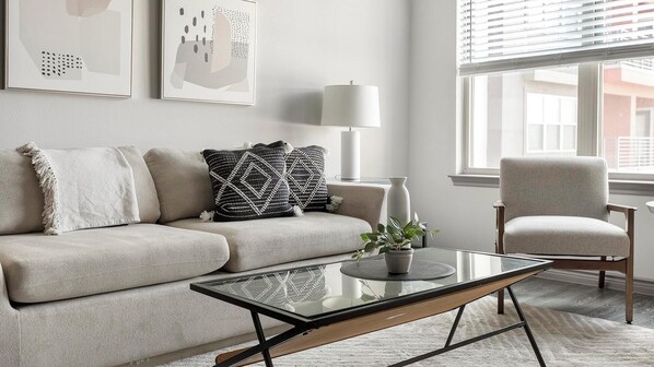 A modern living room with a beige sofa, a glass coffee table, and a grey armchair. There are two black and white patterned throw pillows on the sofa. A potted plant is on the coffee table, and a white floor lamp is beside the sofa. The room has a large window with blinds, and artwork is displayed on the wall.