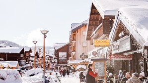 Una calle nevada en un pueblo con edificios de madera, mesas al aire libre en cafés y carteles de tiendas y servicios locales.