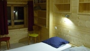 Interior de una cabaña de madera con una cama, una silla y estanterías.