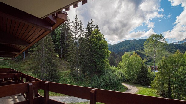 Un balcón de madera con vistas a un paisaje montañoso, un camino de grava y una casa a lo lejos.
