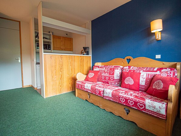 A cozy room with a wooden sofa bed adorned with red and white patterned bedding, a green carpet, a blue accent wall, and a kitchen area with wooden cabinets and a microwave.