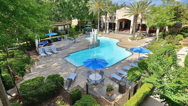 A swimming pool with lounge chairs and umbrellas, surrounded by greenery and buildings. - Landing | Stunning 1BD, Pool, Gym (Houston)