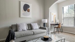 A modern living room with a grey sofa, a glass coffee table, and a desk with a chair. - Landing | Stunning 1BD, Pool, Gym (Houston)