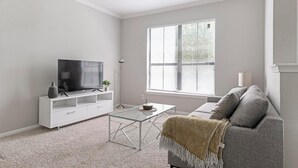 A modern living room with a flat-screen TV on a white TV stand, a glass coffee table, a grey sofa, and a large window with blinds. - Landing | Stunning 1BD, Pool, Gym (Houston)