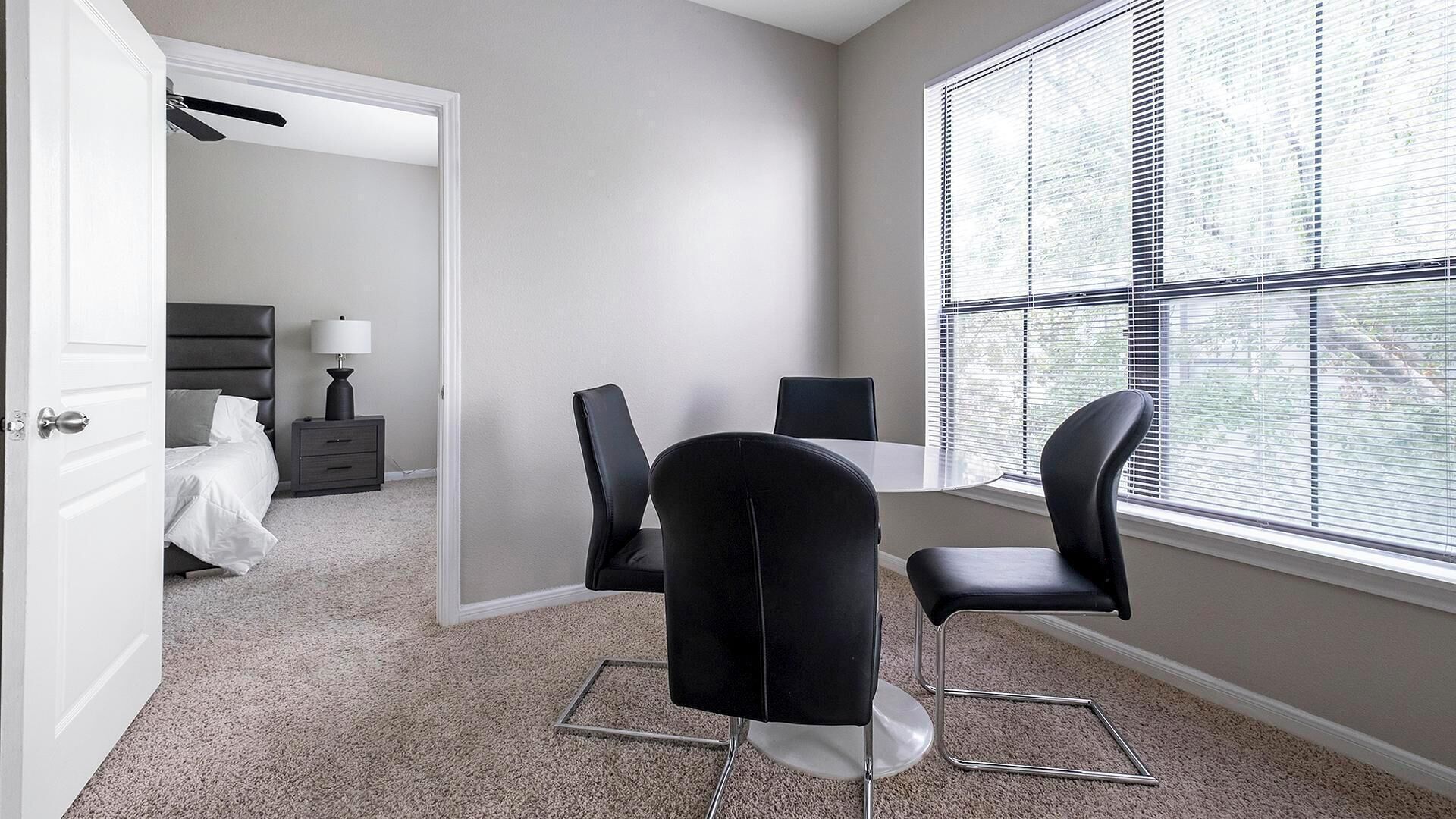 A modern bedroom with a bed, bedside table, and lamp. A dining area with a table and chairs is visible through an open door.