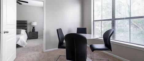 A modern bedroom with a bed, bedside table, and lamp. A dining area with a table and chairs is visible through an open door.