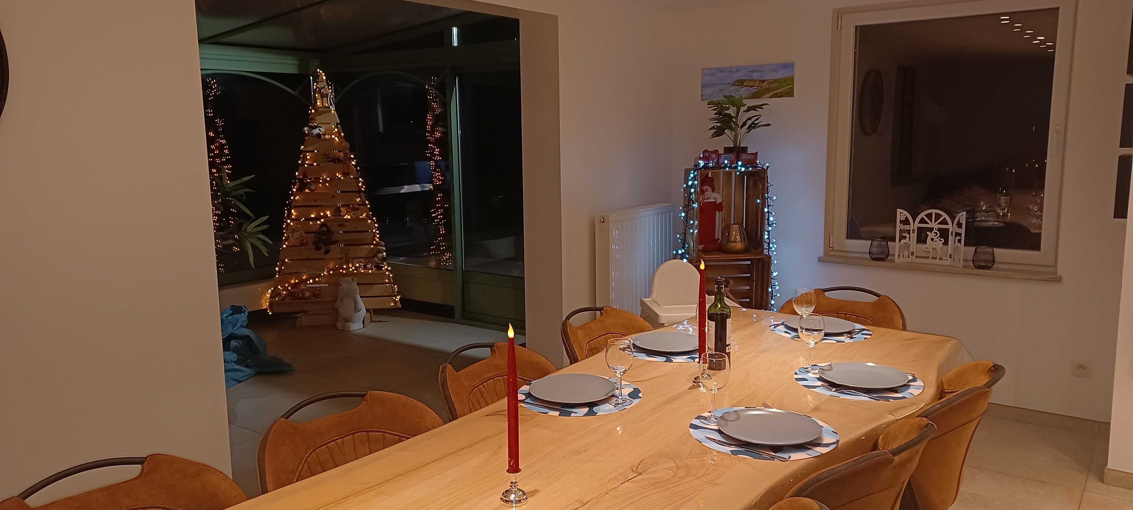 A dining area with a wooden table set for four, a Christmas tree, and a window with a view of the outdoors.
