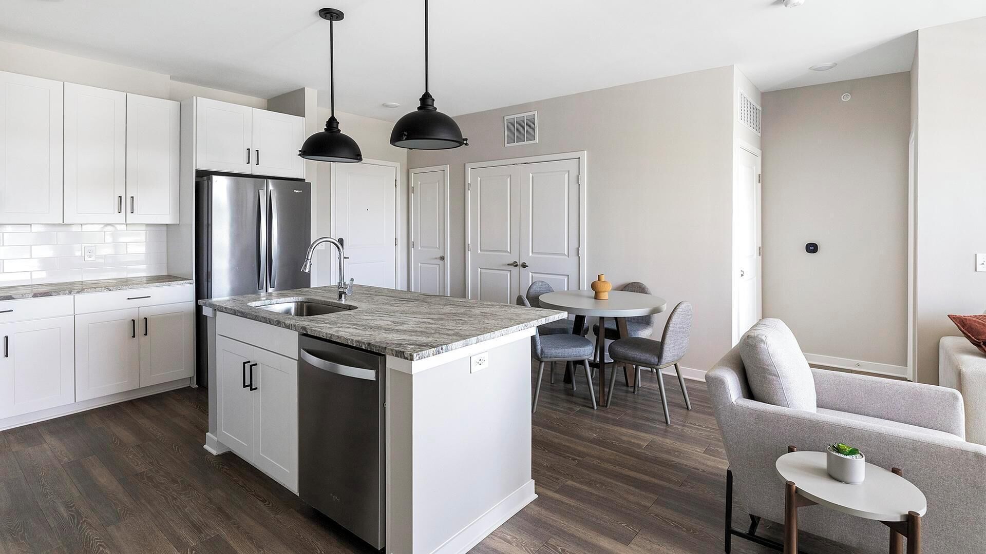 A modern kitchen with white cabinets, a stainless steel refrigerator, a double sink, and a dining area with a round table and chairs.