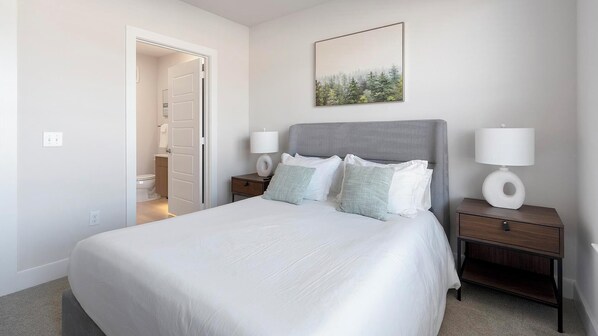 Une chambre avec un grand lit, deux tables de chevet avec des lampes, une porte donnant sur une salle de bain et un tableau encadré au mur.