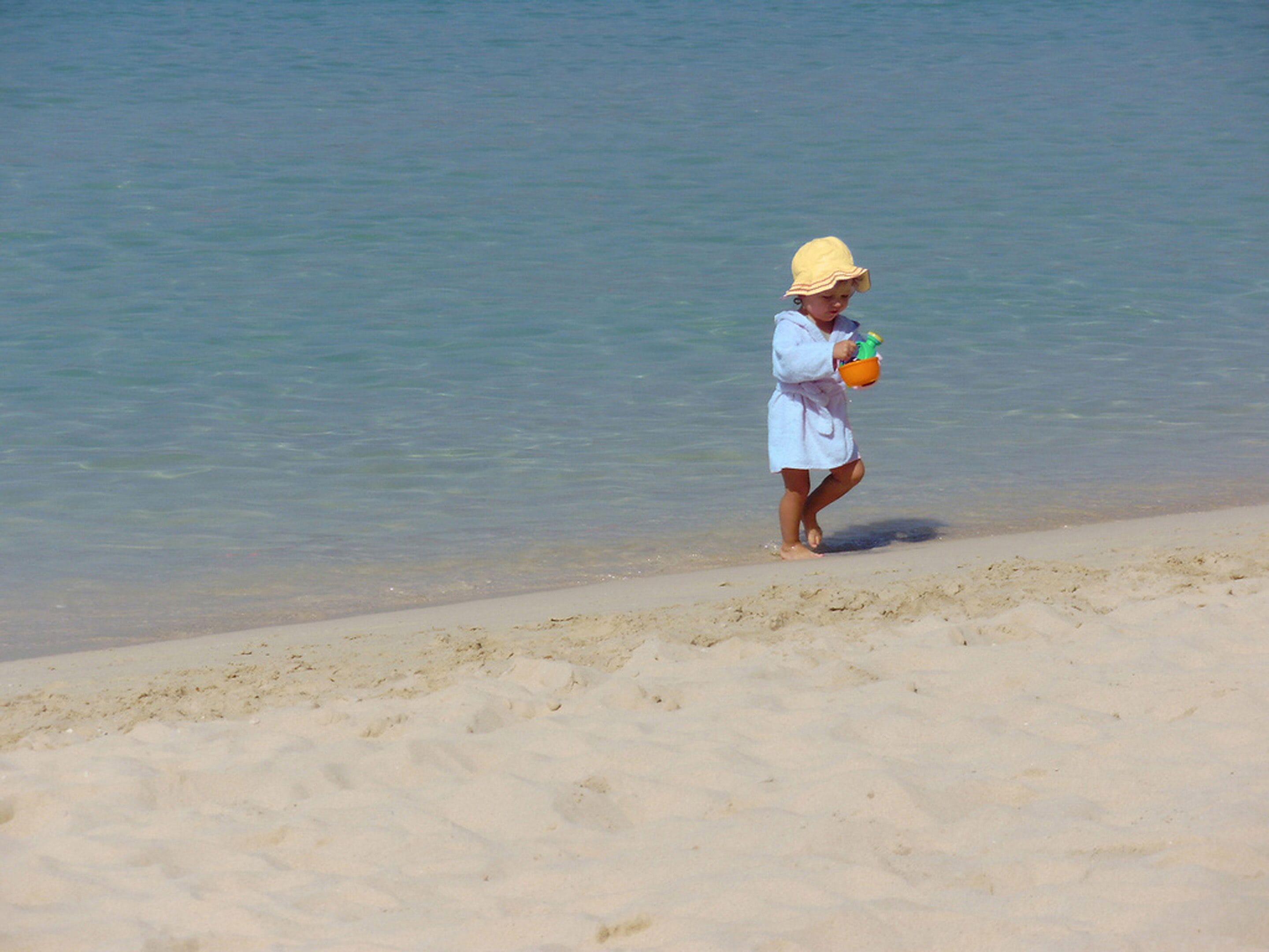 Une personne vêtue d’une robe blanche et d’un chapeau jaune, debout sur une plage de sable près de l’eau.