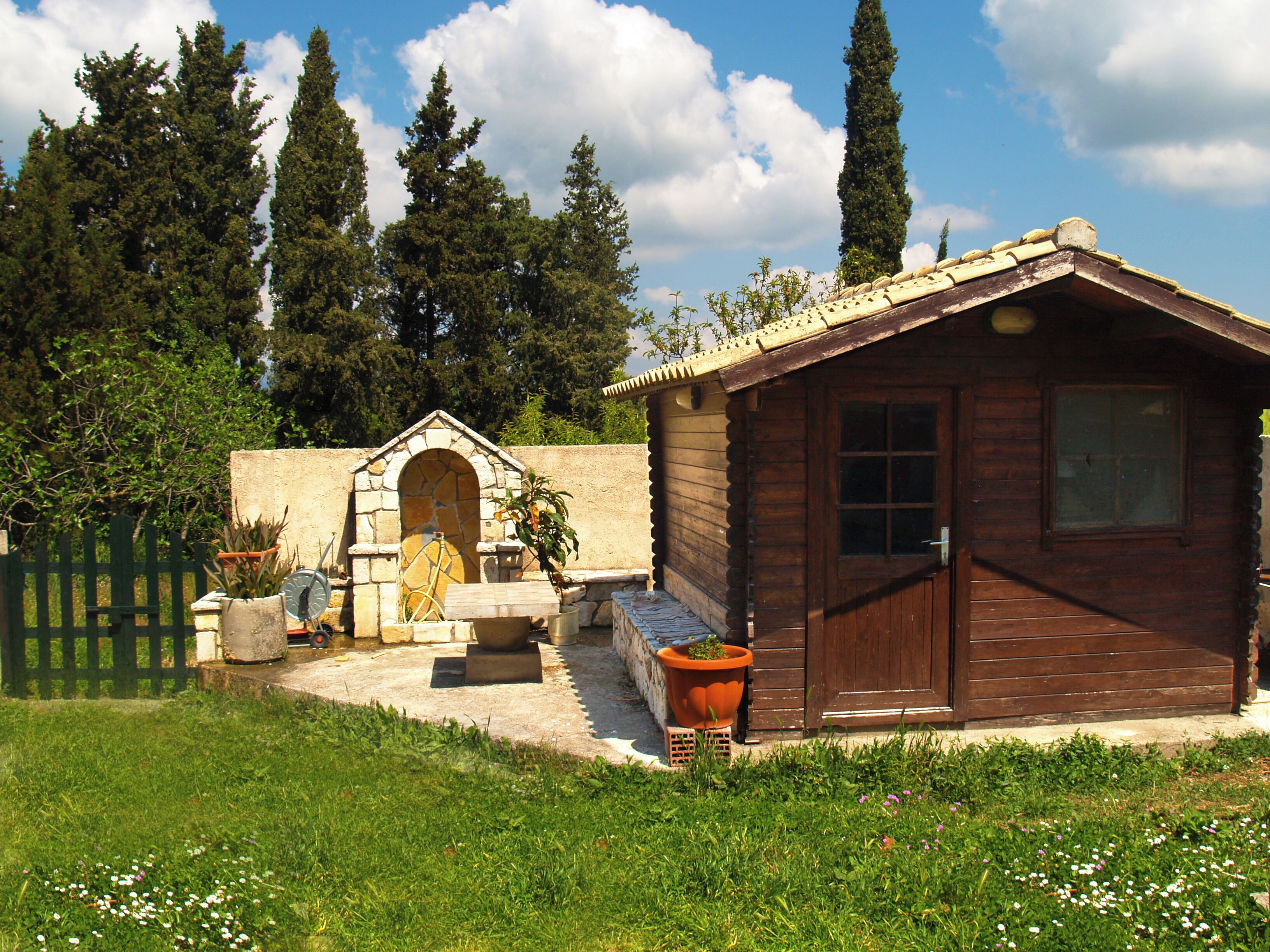 A wooden cabin with a stone archway entrance, surrounded by a garden with potted plants and a wooden fence.