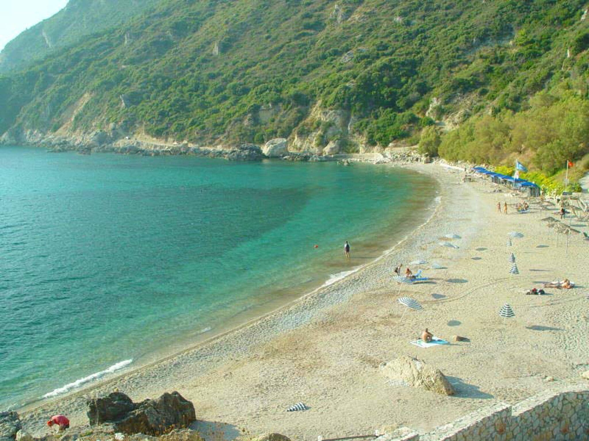 A beach with clear turquoise waters, rocky outcrops, and a few people.
