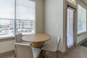 A room with a round wooden table and two chairs, large windows with blinds, and a view of a parking lot.