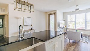 Een moderne keuken met een zwart aanrecht, een roestvrijstalen spoelbak en een hanglamp. Er is een eethoek met een stoel en een tafel, en een raam met zonwering.