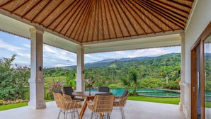 An outdoor covered patio with a table and chairs, overlooking a lush green landscape and a pool.