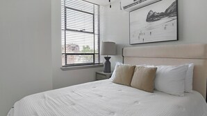 Ein Schlafzimmer mit Bett, Fenster mit Jalousien, Nachttisch mit Lampe und einem gerahmten Bild an der Wand.