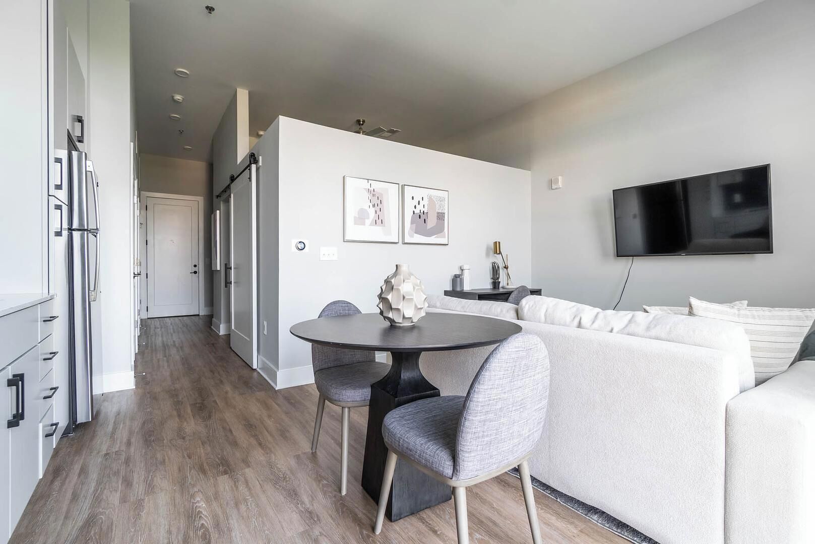 A modern living room with a grey sofa, a dining table with two chairs, and a flat-screen TV mounted on the wall.