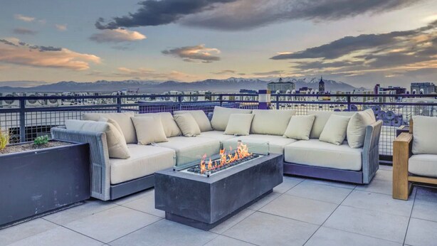 A rooftop terrace with a sectional sofa and a fire pit, overlooking a cityscape and mountains. - Landing | Modern 1BD, Courtyard, Clubhouse (Salt Lake City)