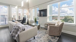 A spacious living room with a large flat-screen TV, comfortable seating, and large windows offering a view of the outdoors.