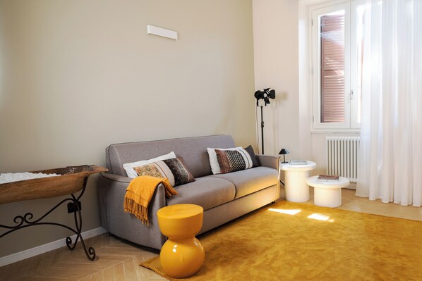 A modern living room with a grey sofa, a wooden side table, a yellow stool, and a radiator near a window with blinds.
