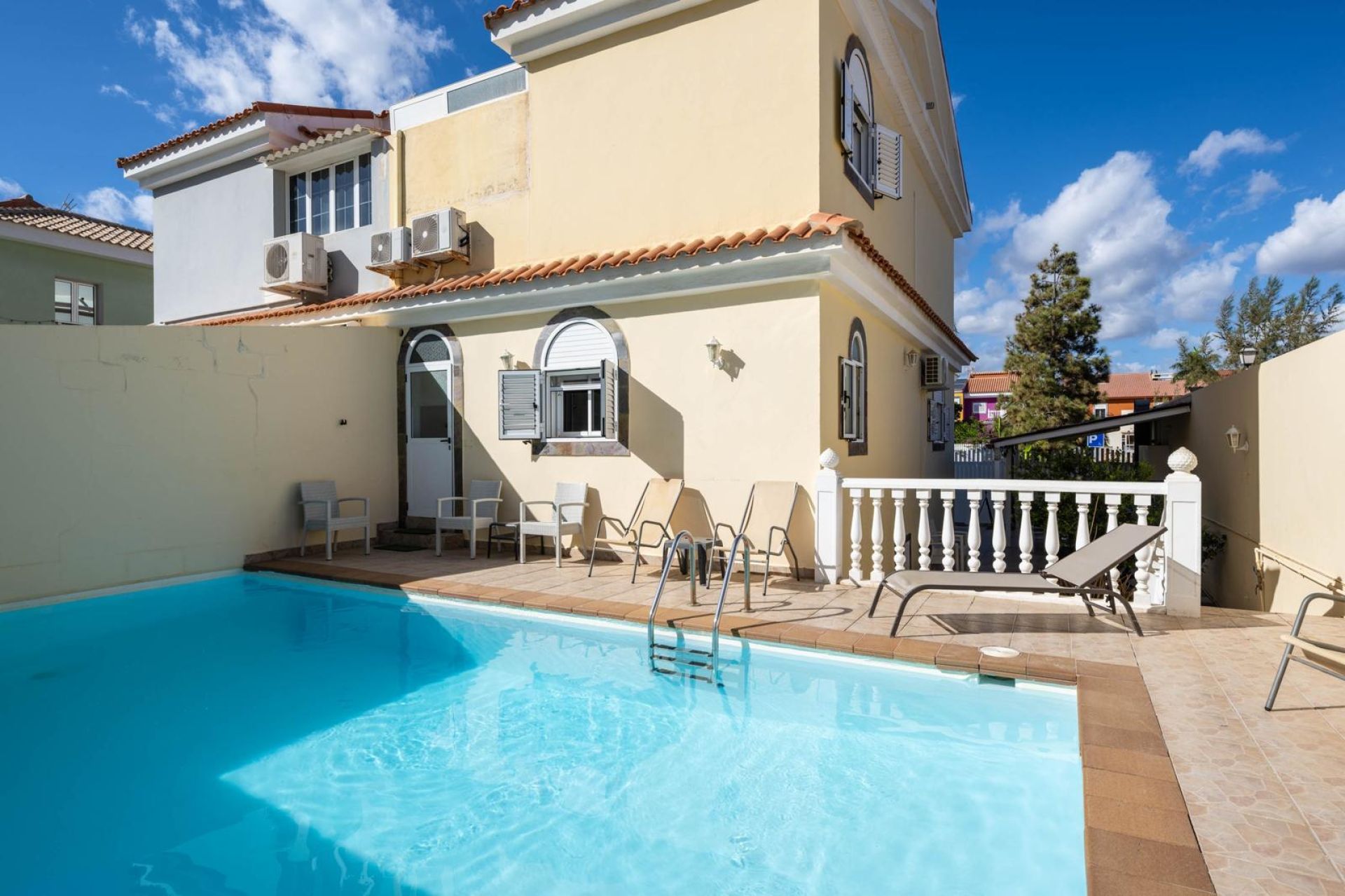 Une piscine avec un solarium, des espaces de détente en plein air et un bâtiment doté d’un toit en tuiles et de climatiseurs.