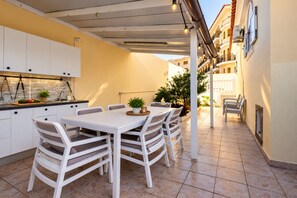 A covered outdoor dining area with a white table and chairs, a kitchen with white cabinets, and a view of a building with balconies.