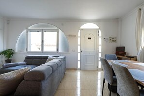 Ein Zimmer mit einer Couch, einem Esstisch mit Stühlen, einem Blumentopf und einer Tür zu einem weiteren Raum.