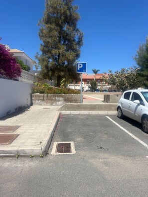 Ein Parkbereich mit einem blauen Schild, das einen Stellplatz kennzeichnet, einem geparkten weißen Auto und einem Gebäude mit rotem Dach im Hintergrund.