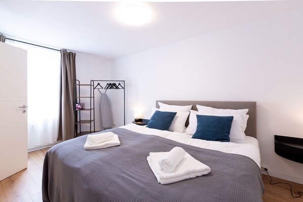 Una habitación de hotel con una cama, dos almohadas, una mesita de noche, un televisor montado en la pared y un perchero con perchas.