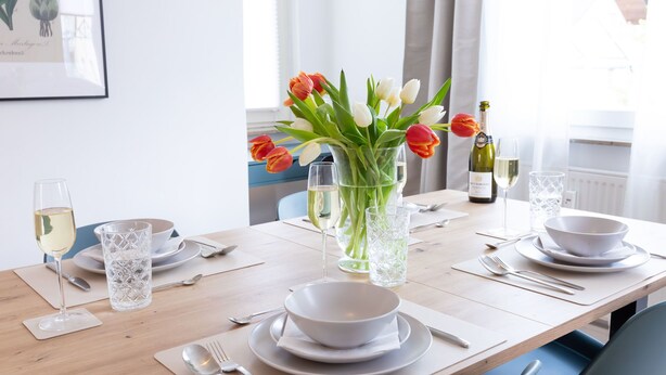 A dining table set with white plates, bowls, cutlery, and glasses, with a vase of tulips and a bottle of wine on the table.
