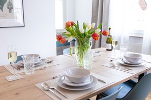 Una mesa de comedor dispuesta con platos, tazones, cubiertos y vasos blancos, con un jarrón de tulipanes y una botella de vino sobre la mesa.