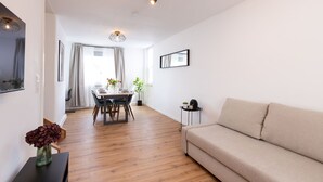 A modern living room with a beige sofa, a dining table with chairs, and a small side table with a vase of flowers.