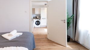 A bedroom with a bed, a washing machine, and a kitchen in the background.