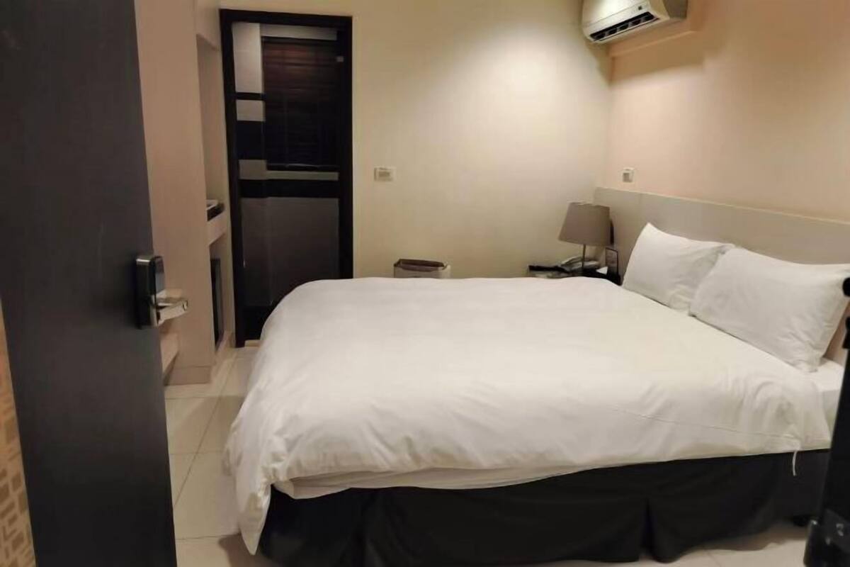 Una habitación de hotel con cama, mesita de noche, lámpara y un equipo de aire acondicionado.