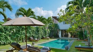 Un espace aménagé au bord de la piscine, avec des chaises longues, un parasol et une maison en arrière-plan.