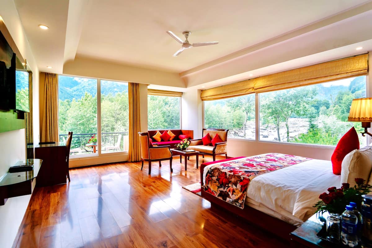 A spacious bedroom with a large bed, a sitting area with a sofa and chairs, a balcony with a view of the mountains, and a ceiling fan.