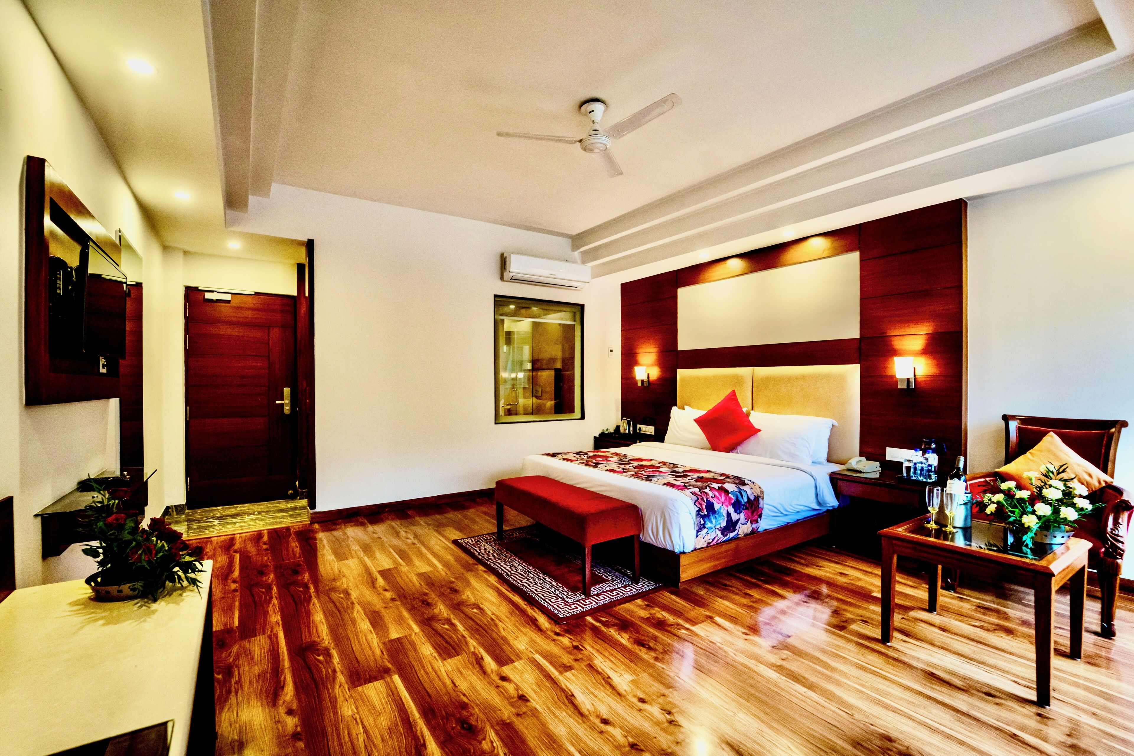 A hotel room with a wooden floor, a bed with red and white bedding, a bedside table with a vase of flowers, a nightstand, and a ceiling fan.