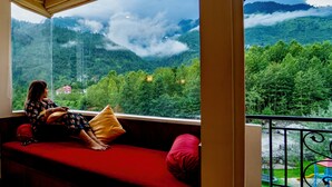 A person sitting on a red sofa with a view of a mountainous landscape through a large window. - The Allure Grand Resort  (Manali)