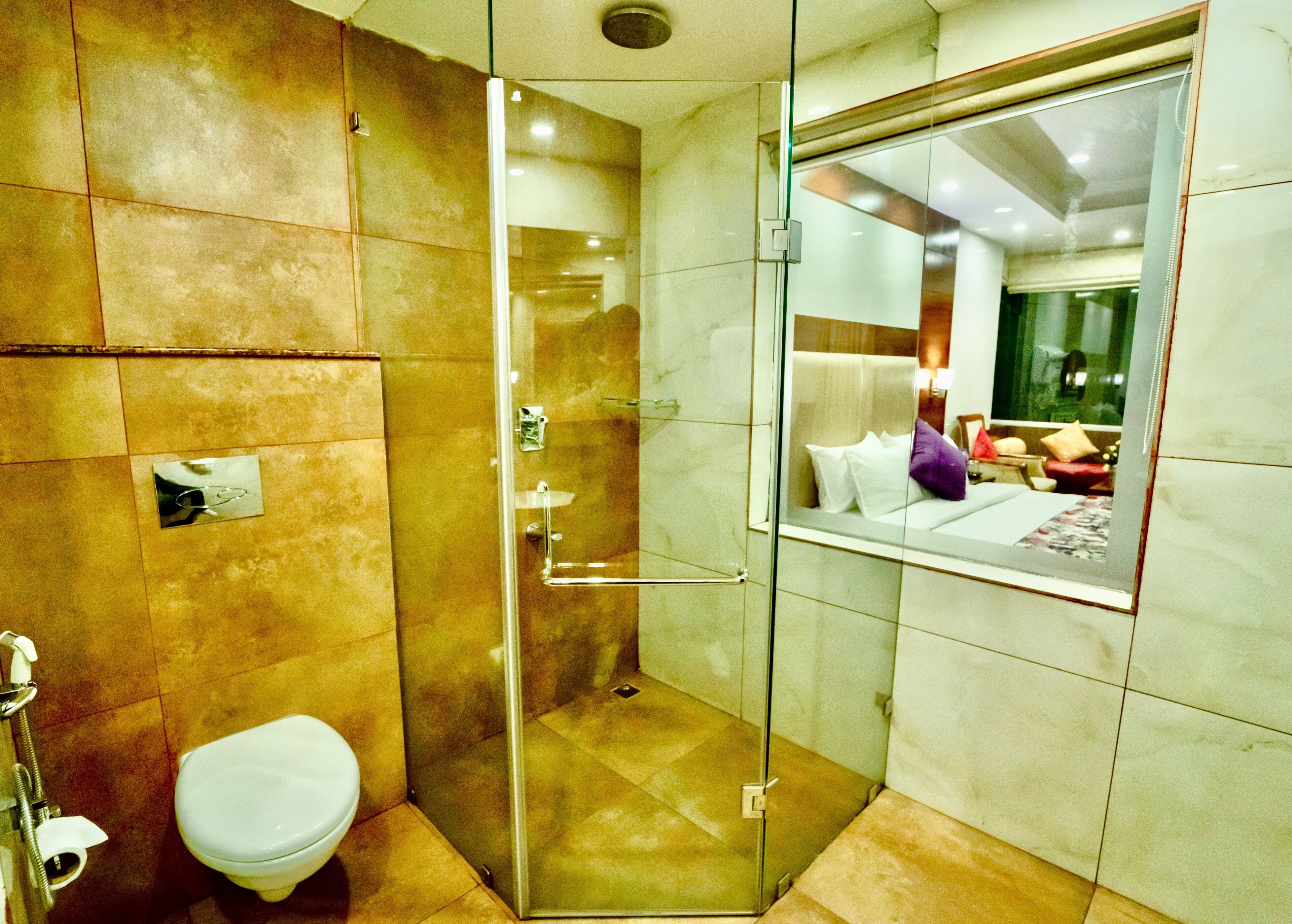 Casa de banho com cabine de chuveiro em vidro, sanita e espelho a refletir uma cama com almofadas.