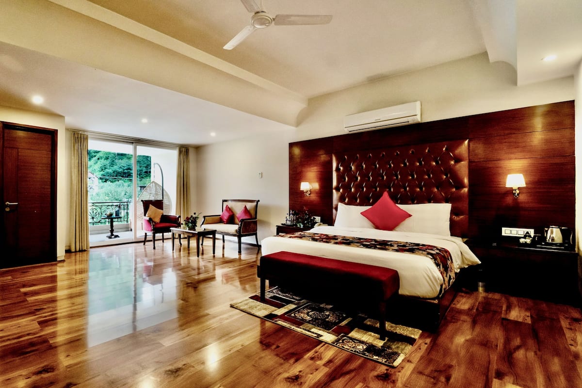 A spacious hotel room with a large bed, a sitting area with a chair and table, a balcony with a view of greenery, and a ceiling fan.