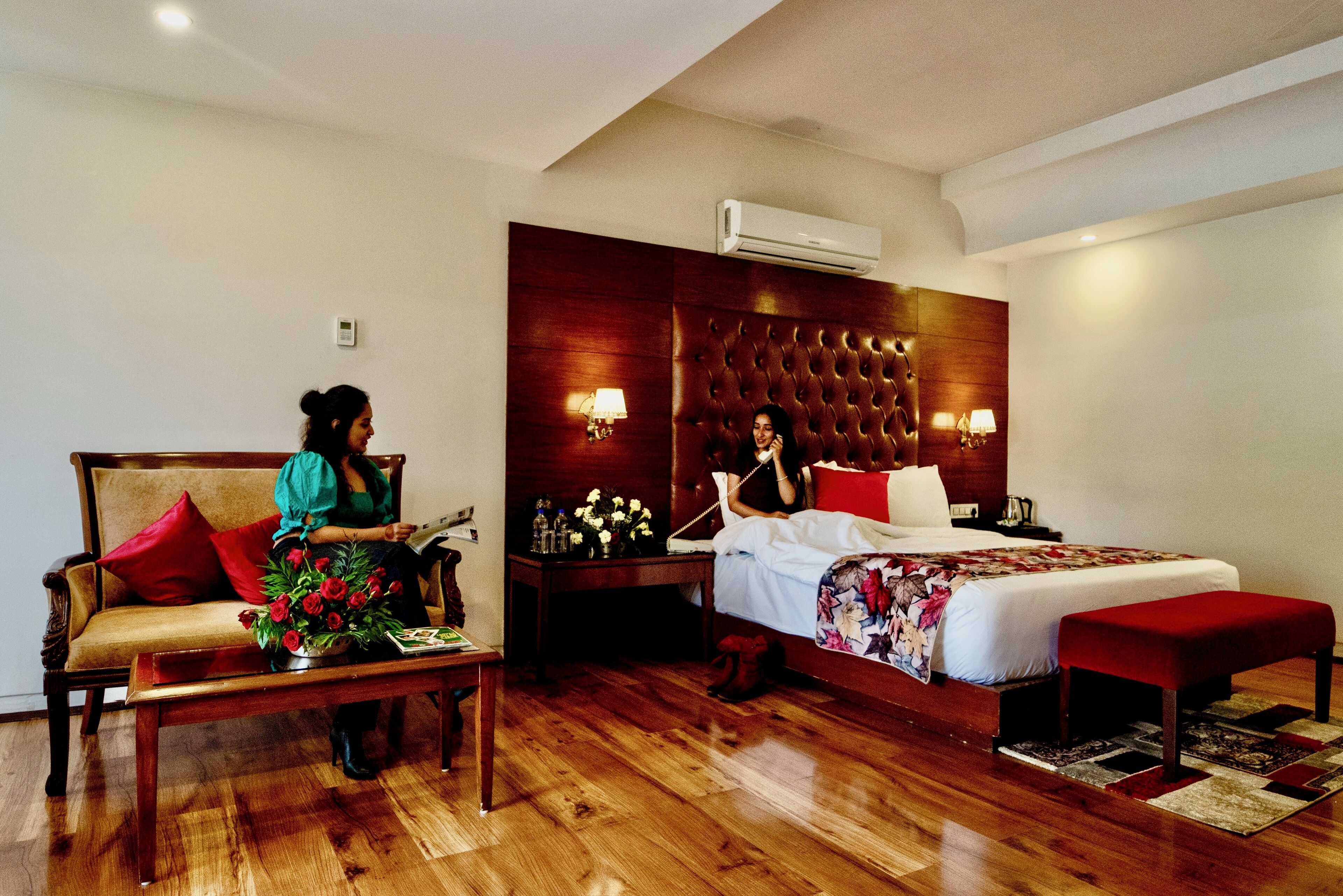 A hotel room with a wooden floor, a sofa with red cushions, a bed with a floral bedspread, and a nightstand with a lamp.